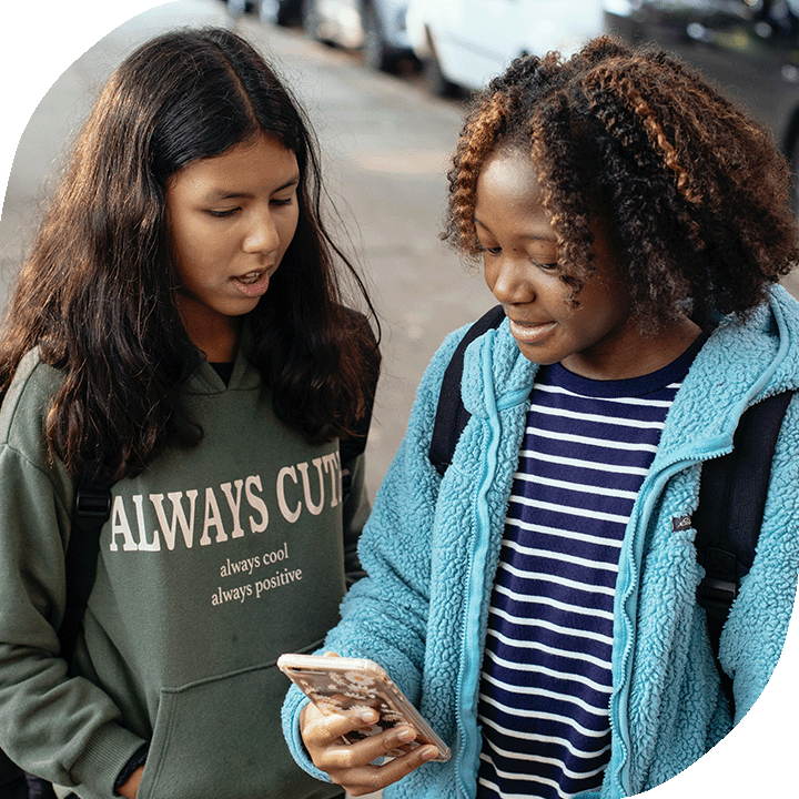 Two young girls are talking about something they see on one girl's phone. Both are school-aged and wearing hoodies and backpacks. One has medium tone skin and long brown hair and the other is Black with medium-length curly hair.