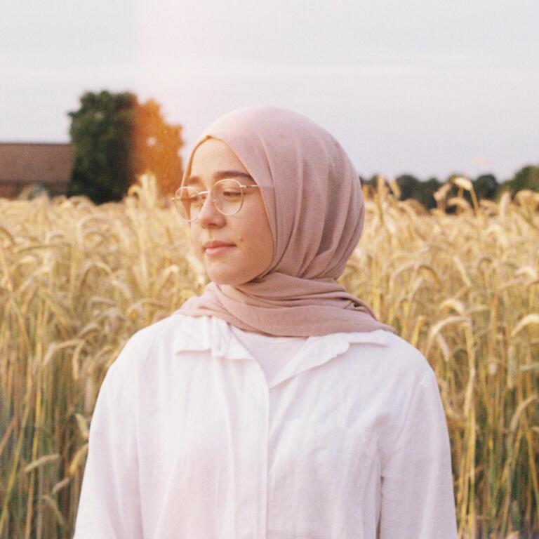 Young woman wearing a light pink hijab and glasses stands in front of a wheat field.
